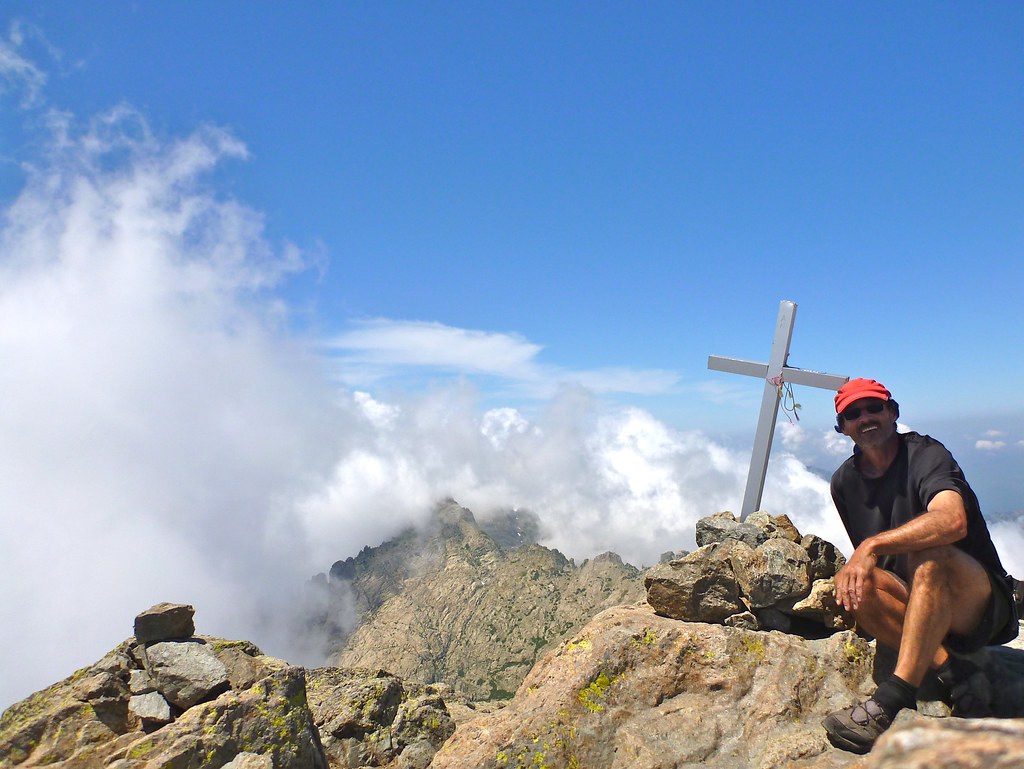 Un randonneur à la croix sommitale du Monte d'Oro sur le GR20, nuages en contrebas